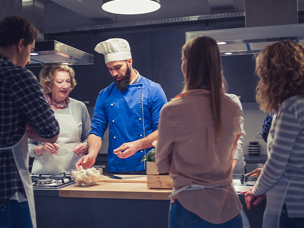Szene Küche: Mehrere Personen stehen um den Koch herum und schauen seinen Anweisungen zu